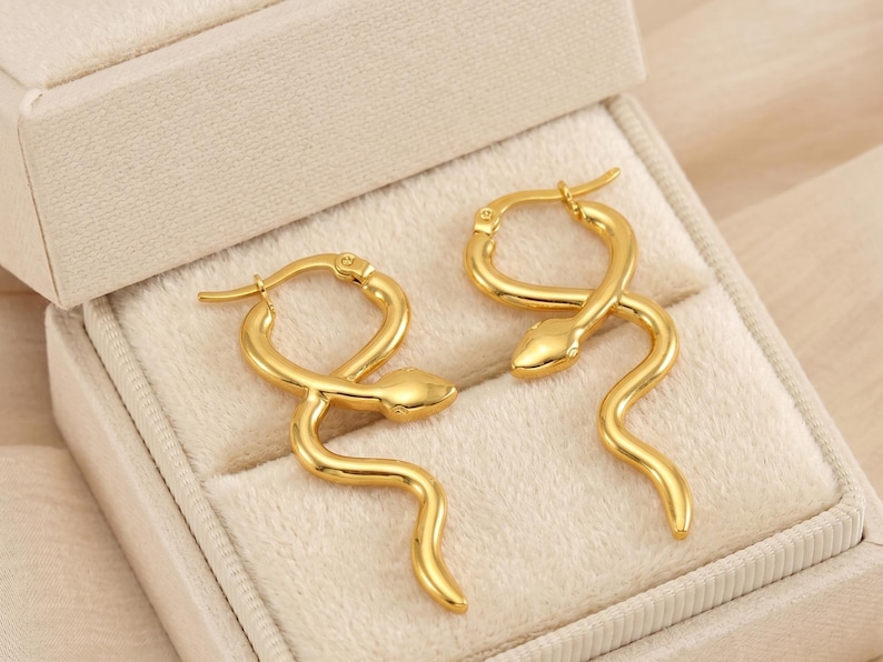 May include: A pair of gold-colored snake-shaped earrings. The earrings are in a box with a cream-colored interior. The earrings have a hoop design and a snake head at the top. The snake's body curves downward.