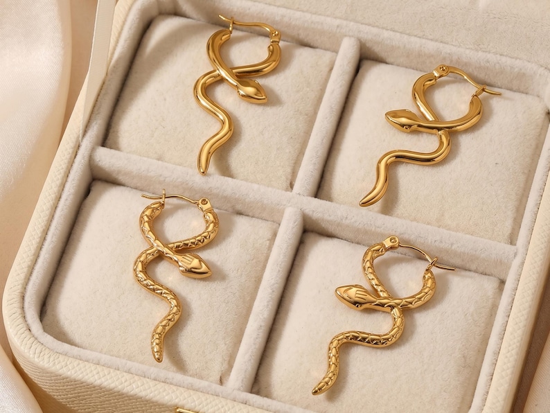 May include: Gold-colored snake-shaped hoop earrings displayed in a jewelry box. The earrings have a detailed snake design, with the snake's head and body forming the hoop. The jewelry box has a neutral-colored interior.
