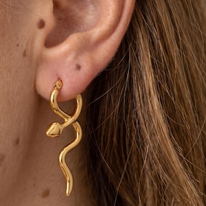 May include: Gold-colored hoop earrings with a snake design. The earrings feature a coiled snake shape, with the head at the top and the body curving down. The earrings are made of a shiny, reflective metal.