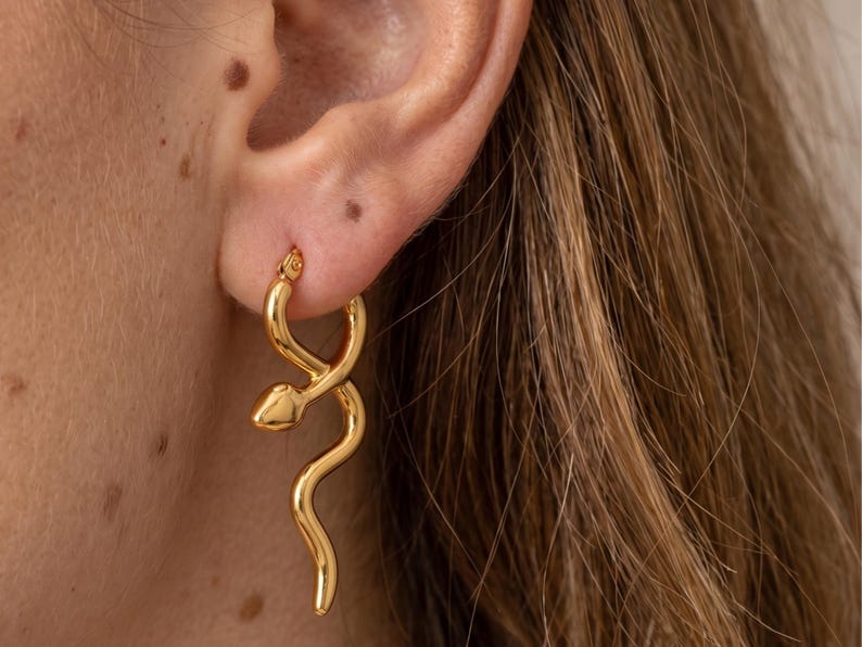 May include: Gold-colored hoop earrings with a snake design. The earrings feature a coiled snake shape, with the head at the top and the body curving down. The earrings are made of a shiny, reflective metal.