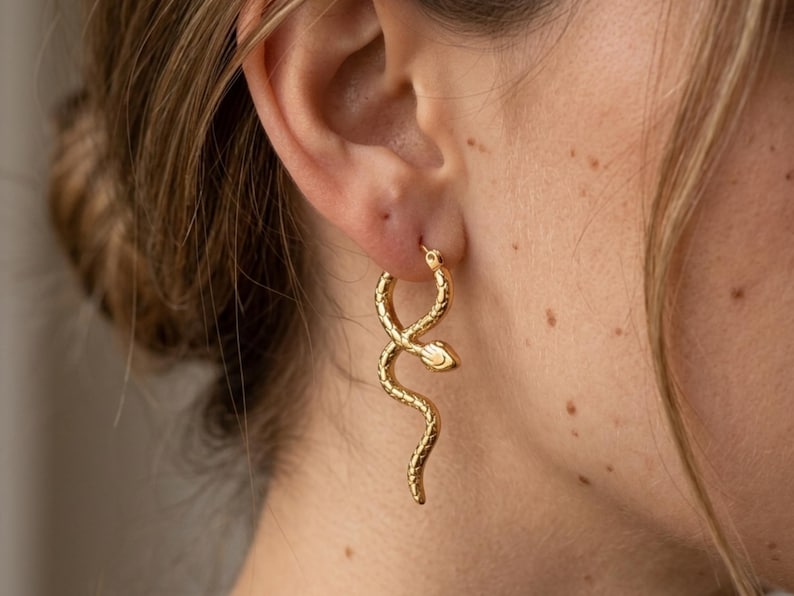 May include: Gold-colored snake hoop earrings. The earrings are worn on the ear and feature a detailed snake design. The snake's body curves and twists, with a small snake head at the bottom of the hoop. The earrings are a stylish accessory.