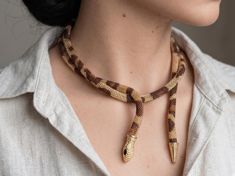 May include: A gold and brown snake necklace with a detailed design. The necklace is wrapped around the neck and features a snake head with a black eye and a pointed tail. The necklace is made of small, woven beads.