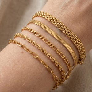 May include: Several gold bracelets of different chain designs, including a herringbone, rope, and link style. The bracelets are stacked on a wrist, displaying the various textures and patterns of the jewellery.