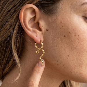 May include: Gold-colored snake-shaped hoop earrings. The earrings are worn on a person's ear. The snake design features a curved body and a small head. The earrings are made of a shiny metal and are a stylish accessory.