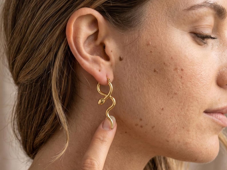 May include: Gold-colored snake-shaped hoop earrings. The earrings are worn on a person's ear. The snake design features a curved body and a small head. The earrings are made of a shiny metal and are a stylish accessory.