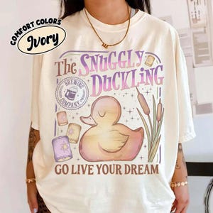 May include: Ivory Comfort Colors t-shirt featuring a whimsical design. The graphic includes a duck, the text "The Snuggly Duckling Brewing Company", and the phrase "Go Live Your Dream". The design is in shades of pink, purple, and yellow.