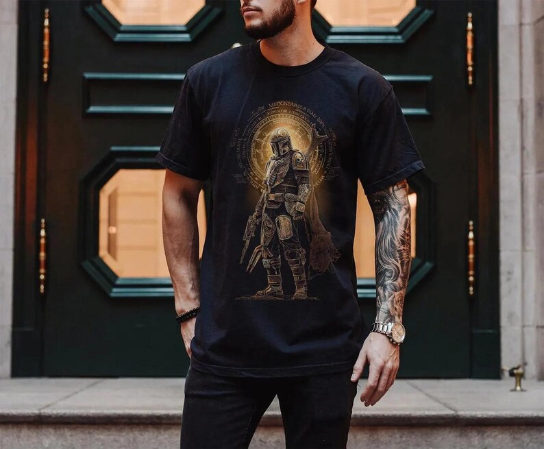 May include: A black short-sleeved t-shirt featuring a graphic print of a armored character in a helmet standing in front of a glowing golden circular emblem. The design includes intricate line work and a weathered, metallic aesthetic.
