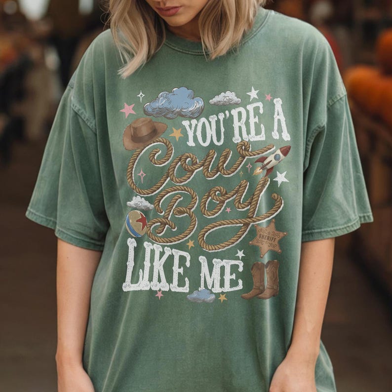 K&ouml;nnte beinhalten: Ein salbeigr&uuml;nes Kurzarm-T-Shirt mit einem Grafikdruck und dem Text "YOU'RE A COWBOY LIKE ME" in wei&szlig;er Schrift. Das Design enth&auml;lt Illustrationen eines braunen Cowboyhuts, eines Seils, einer Rakete, eines Balls, eines Sheriffsterns und Stiefeln.