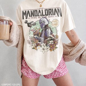 Op de afbeelding: Ivoor T-shirt met een afbeelding van The Mandalorian en Grogu, omringd door bloemen, met de tekst "THE MANDALORIAN THIS IS THE WAY". Het shirt wordt gecombineerd met een roze en witte geruite short.