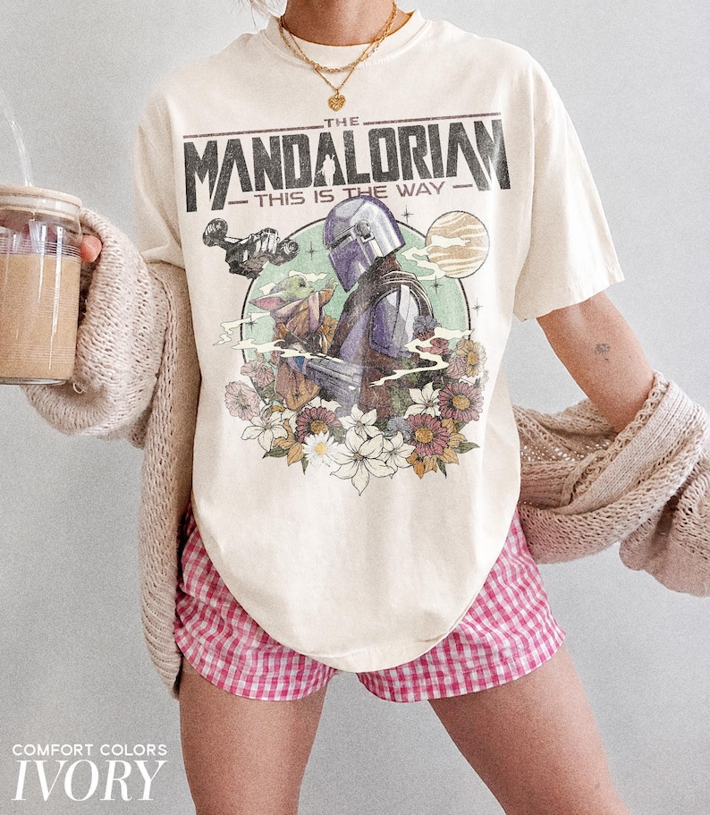 Op de afbeelding: Ivoor T-shirt met een afbeelding van The Mandalorian en Grogu, omringd door bloemen, met de tekst "THE MANDALORIAN THIS IS THE WAY". Het shirt wordt gecombineerd met een roze en witte geruite short.