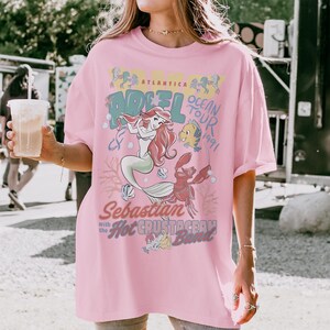 May include: A light pink t-shirt featuring a graphic print of Ariel, Flounder, and Sebastian from The Little Mermaid. The text reads Atlantica Ocean Tour 1991, Sebastian with the Hot Crustacean Band, and Ariel. The design includes coral and shell motifs.