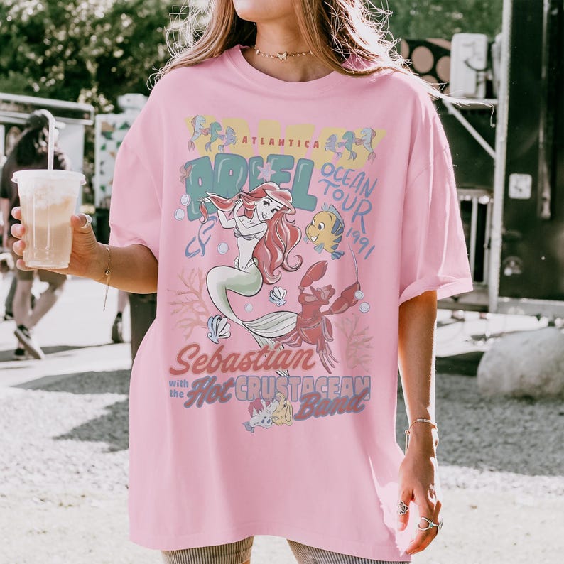 May include: A light pink t-shirt featuring a graphic print of Ariel, Flounder, and Sebastian from The Little Mermaid. The text reads Atlantica Ocean Tour 1991, Sebastian with the Hot Crustacean Band, and Ariel. The design includes coral and shell motifs.
