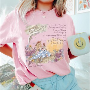 Puede incluir: Una camiseta rosa claro con una ilustraci&oacute;n gr&aacute;fica de una persona con sombrero de vaquero y vestido azul sentada en un campo de flores. El dise&ntilde;o incluye la letra impresa: I remembered I loved you, Come back when it mattered, I saw you standing there in the light of the window wearing that same smile, Man, it's been a while, But I knew it, I knew you, I knew it, I knew you, I knew it, I knew you.