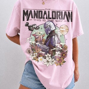 Op de afbeelding: Roze T-shirt met een afbeelding van The Mandalorian, Grogu en een ruimteschip, omringd door bloemen. De tekst "THE MANDALORIAN" en "THIS IS THE WAY" staat boven de afbeelding. Het shirt is gelabeld "COMFORT COLORS BLOSSOM".