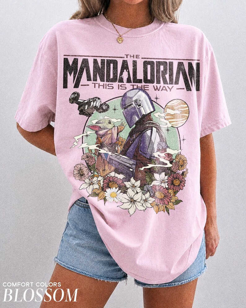 Op de afbeelding: Roze T-shirt met een afbeelding van The Mandalorian, Grogu en een ruimteschip, omringd door bloemen. De tekst "THE MANDALORIAN" en "THIS IS THE WAY" staat boven de afbeelding. Het shirt is gelabeld "COMFORT COLORS BLOSSOM".