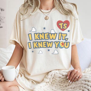 Op de afbeelding: Een cr&egrave;mekleurig t-shirt met korte mouwen en de gele tekst I KNEW IT, I KNEW YOU met blauwe omlijning. Het ontwerp bevat een rood hart met de letters TS, kleine blauwe sterren, witte wolkenomtrekken en een kleurrijke balafbeelding.
