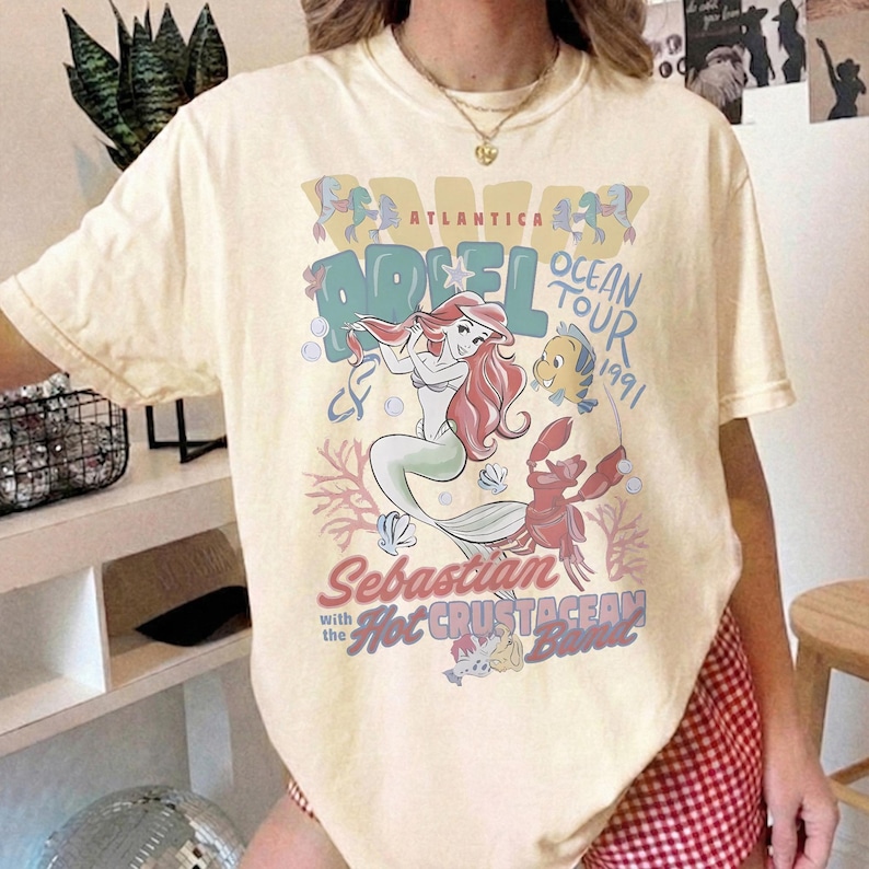 May include: A cream-coloured short-sleeve t-shirt featuring a graphic print of Ariel, Flounder, and Sebastian. The text reads Atlantica Ocean Tour 1991, Sebastian with the Hot Crustacean Band. The design includes colourful illustrations of sea life.