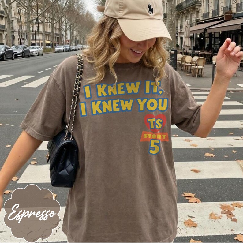 May include: A brown short-sleeved t-shirt featuring the yellow and blue text I KNEW IT, I KNEW YOU, a red heart with TS, and the word STORY above the number 5 in yellow and blue. The shirt is paired with a beige baseball cap and a black handbag.