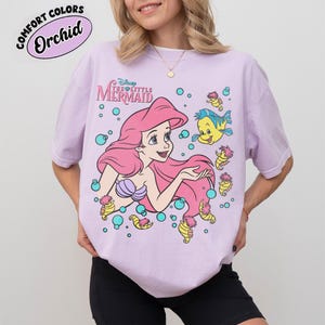 Puede incluir: Una camiseta de color morado claro con un gr&aacute;fico de Ariel de La Sirenita junto a Flounder y caballitos de mar rodeados de burbujas azul verdoso. El texto dice Disney The Little Mermaid. Una etiqueta Comfort Colors Orchid es visible.
