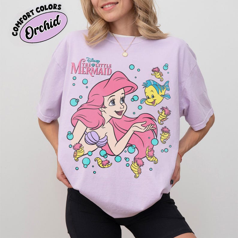 Puede incluir: Una camiseta de color morado claro con un gr&aacute;fico de Ariel de La Sirenita junto a Flounder y caballitos de mar rodeados de burbujas azul verdoso. El texto dice Disney The Little Mermaid. Una etiqueta Comfort Colors Orchid es visible.