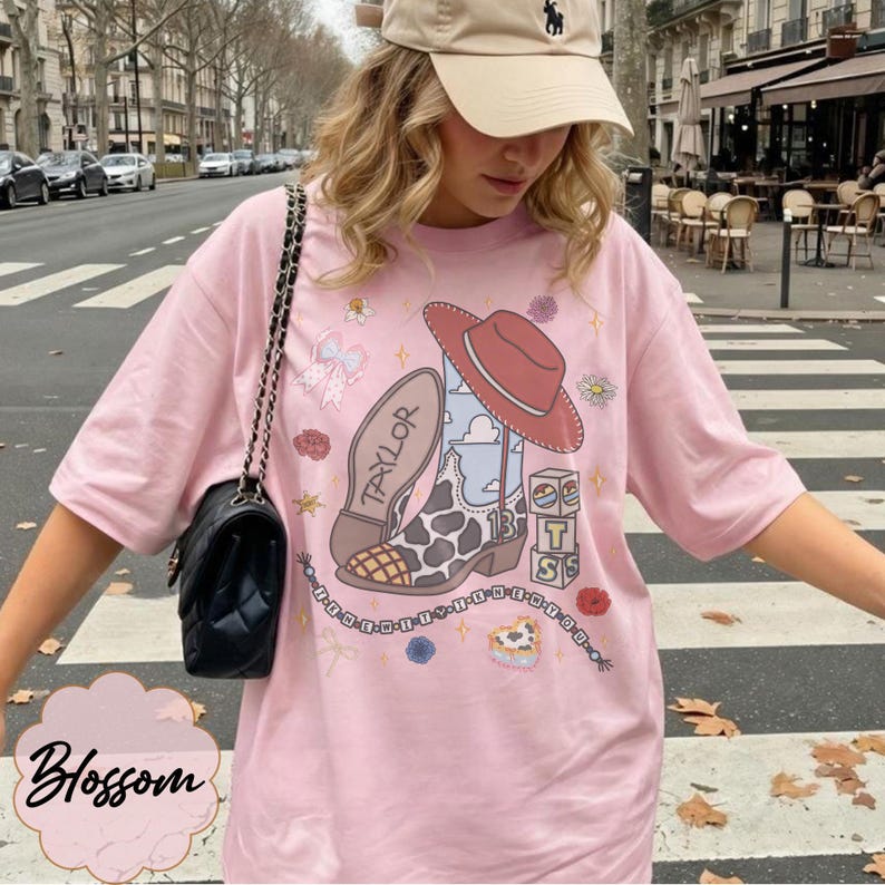Puede incluir: Una camiseta rosa claro con un estampado gr&aacute;fico de una bota vaquera con patr&oacute;n de manchas de vaca, un sombrero rojo y bloques con las letras T, S y el n&uacute;mero 13. El dise&ntilde;o incluye el nombre Taylor, una cadena de cuentas y flores.