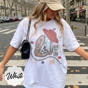 Puede incluir: Una camiseta blanca con un estampado gr&aacute;fico de una bota de vaquero con manchas de vaca, un sombrero rojo, una pila de bloques con las letras T, S, el n&uacute;mero 13 y el nombre Taylor. El dise&ntilde;o incluye peque&ntilde;os detalles florales, estrellas y una cadena.