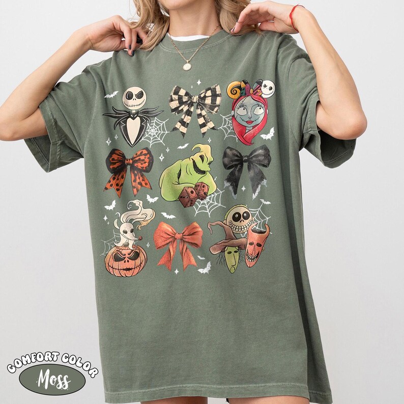 Op de afbeelding: Een olijfgroen T-shirt met korte mouwen en een grafische print van diverse personages en strikken, waaronder een skelet, een groen wezen met dobbelstenen, een spookhond op een pompoen en drie ondeugende figuren, allemaal geaccentueerd met spinnenwebben.