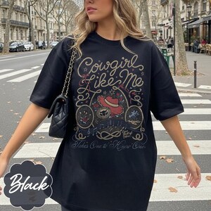 May include: A black short-sleeved t-shirt featuring a graphic print with the text Cowgirl Like Me and Takes One to Know One. The design includes a red cowboy hat, a horseshoe, a rope border, musical notes, and small star accents.
