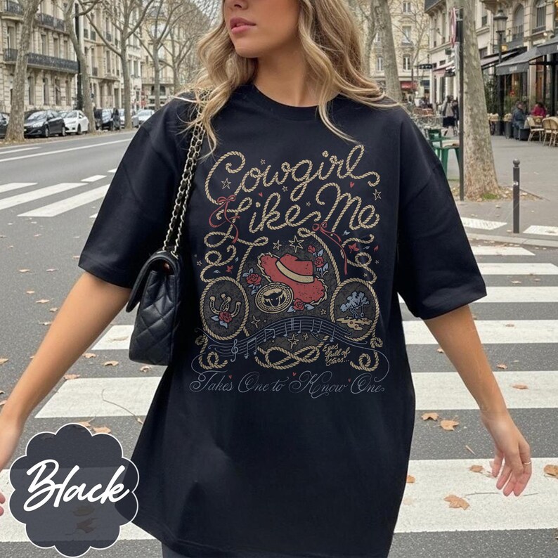 May include: A black short-sleeved t-shirt featuring a graphic print with the text Cowgirl Like Me and Takes One to Know One. The design includes a red cowboy hat, a horseshoe, a rope border, musical notes, and small star accents.