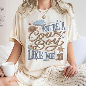 K&ouml;nnte beinhalten: Ein cremefarbenes Kurzarm-T-Shirt mit einem Grafikdruck, der den Text "YOU'RE A COWBOY LIKE ME" in blauen Buchstaben zeigt. Das Design enth&auml;lt Illustrationen eines braunen Cowboyhutes, eines Seils, einer Rakete, eines Sheriffsterns und Stiefeln.