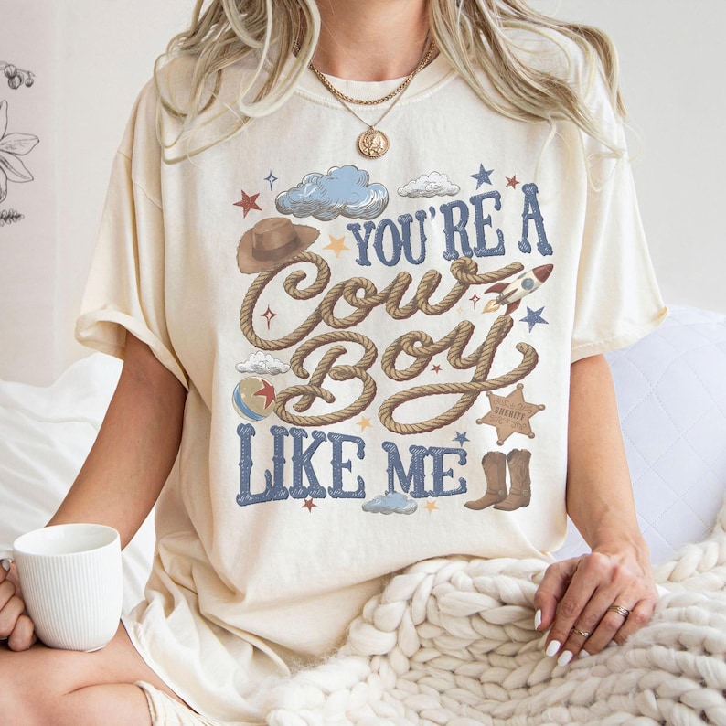 K&ouml;nnte beinhalten: Ein cremefarbenes Kurzarm-T-Shirt mit einem Grafikdruck, der den Text "YOU'RE A COWBOY LIKE ME" in blauen Buchstaben zeigt. Das Design enth&auml;lt Illustrationen eines braunen Cowboyhutes, eines Seils, einer Rakete, eines Sheriffsterns und Stiefeln.