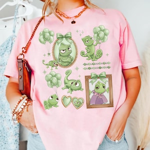 May include: Pink t-shirt featuring a green chameleon character design. The design includes various illustrations of the chameleon with balloons, bows, and framed portraits. The overall theme is a cartoon or animated character.