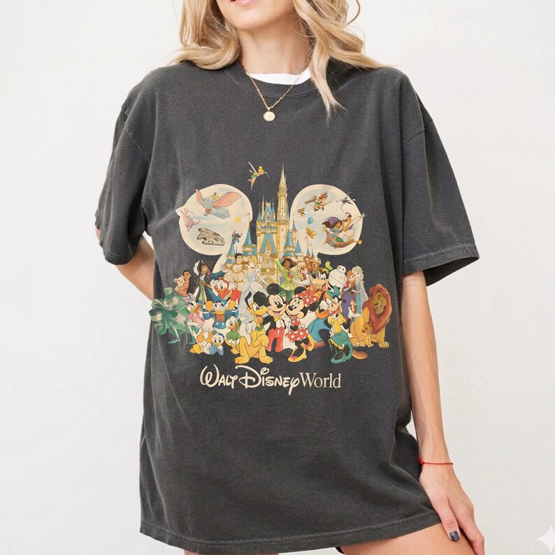 May include: A charcoal grey short-sleeve t-shirt featuring a large graphic print of various Disney characters and a castle silhouette arranged in a Mickey Mouse ear shape. The text Walt Disney World is printed in white below the graphic.