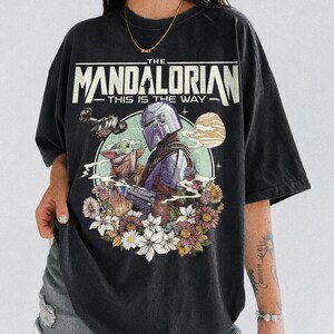 Op de afbeelding: Zwart T-shirt met een afbeelding van The Mandalorian en Grogu, omringd door bloemen. De tekst "THE MANDALORIAN - THIS IS THE WAY" staat bovenaan gedrukt. Het shirt is van het merk Comfort Colors.