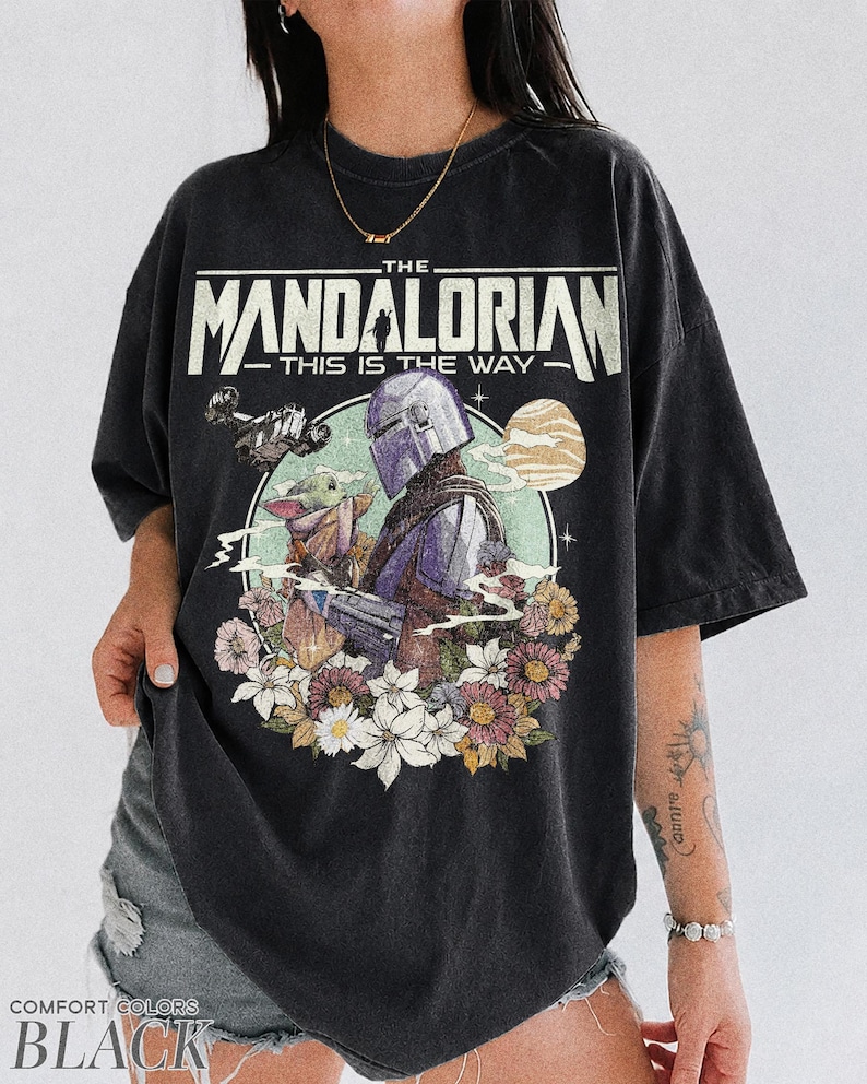 Op de afbeelding: Zwart T-shirt met een afbeelding van The Mandalorian en Grogu, omringd door bloemen. De tekst "THE MANDALORIAN - THIS IS THE WAY" staat bovenaan gedrukt. Het shirt is van het merk Comfort Colors.