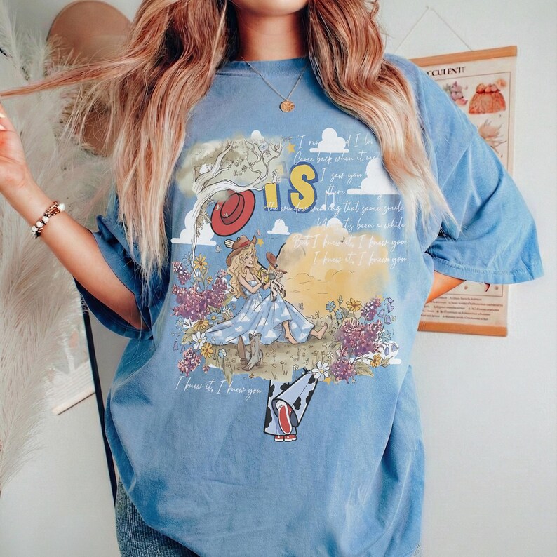 Puede incluir: Una camiseta azul de manga corta con un estampado gr&aacute;fico de un personaje rubio con vestido azul y sombrero de vaquero sentado en un campo de flores. El dise&ntilde;o incluye un sombrero rojo, nubes y el texto I knew it, I knew you.