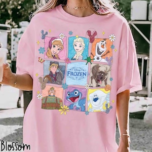 May include: A light pink t-shirt featuring a grid of colorful illustrations of characters from the Disney Frozen movie, including Anna, Elsa, Olaf, Kristoff, Sven, and Bruni, with the Disney Frozen logo in the center and small flower accents.