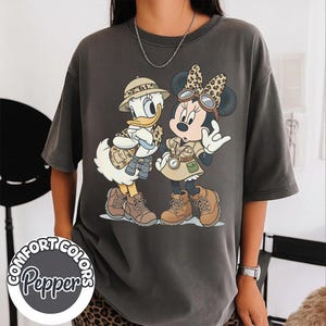 Könnte beinhalten: Dunkelgraues Comfort Colors T-Shirt mit einer Cartoon-Illustration von Donald Duck und Minnie Mouse in Safari-Outfits. Das Shirt hat die Worte "Comfort Colors Pepper" in einem kreisförmigen Design.