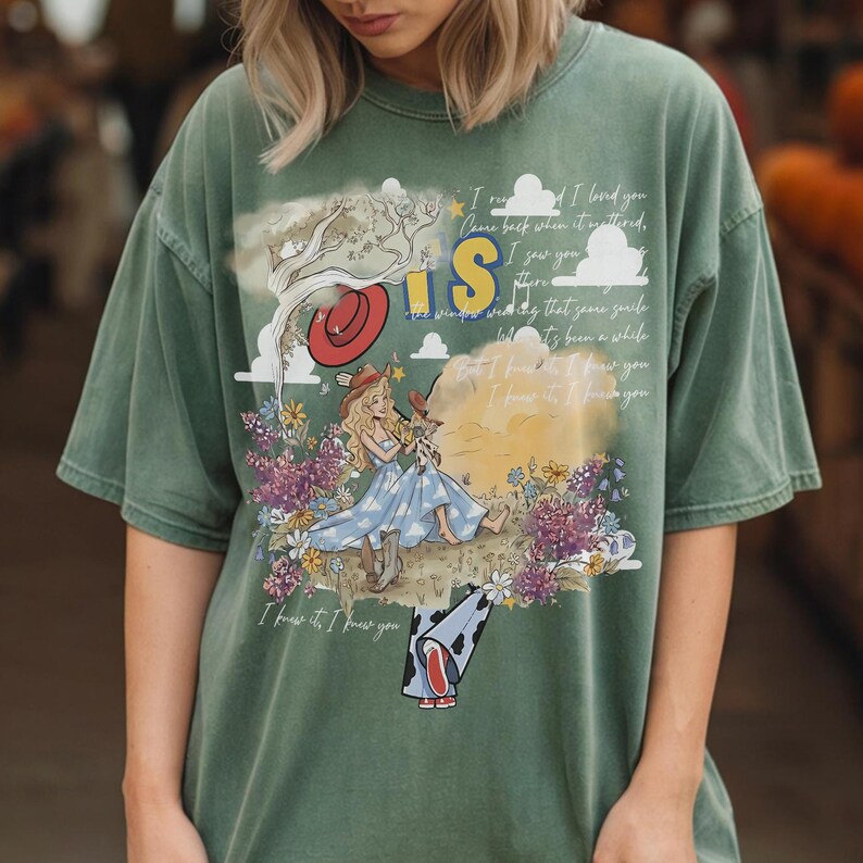 Puede incluir: Una camiseta de manga corta color verde salvia con un estampado gr&aacute;fico de un personaje rubio con vestido azul y sombrero de vaquero sentado en un campo de flores. El dise&ntilde;o incluye un sombrero rojo, nubes, un &aacute;rbol y texto cursivo.