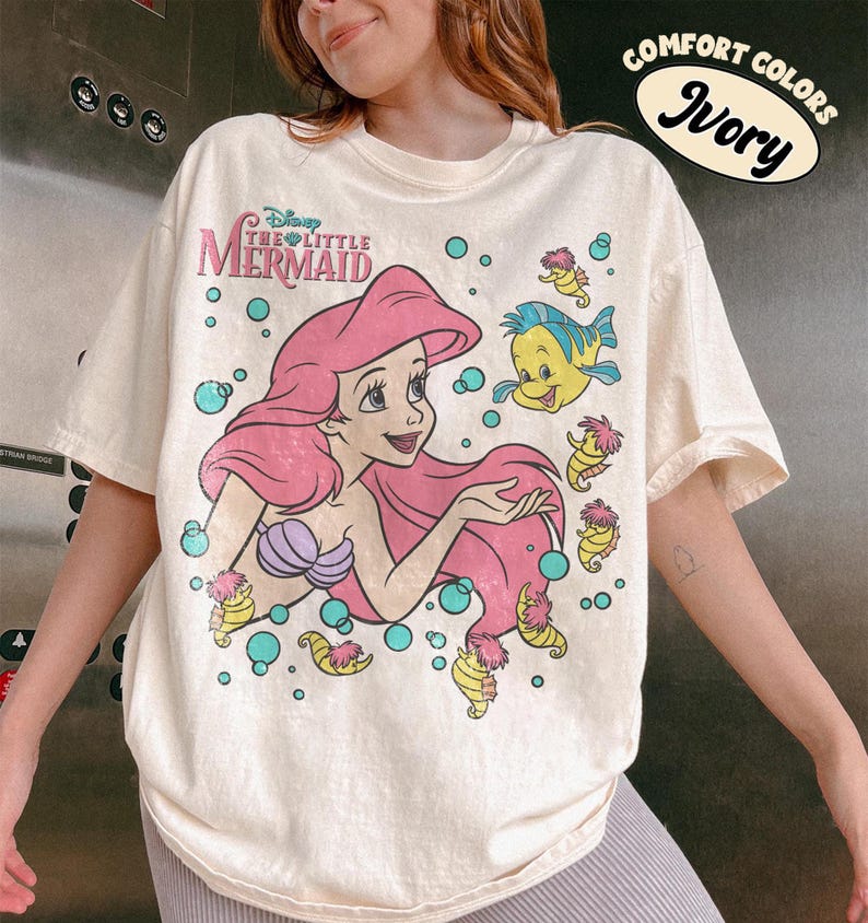 Puede incluir: Una camiseta de color marfil con un estampado gr&aacute;fico de Ariel de La Sirenita con pelo rosa, un top de conchas morado y Flounder. El dise&ntilde;o incluye caballitos de mar amarillos y burbujas verde azulado. Una pegatina en la esquina dice Comfort Colors Ivory.