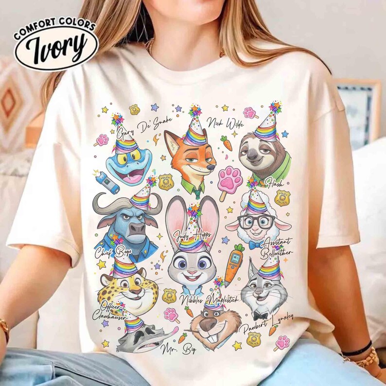 Op de afbeelding: Ivoor kleurig t-shirt met cartoonfiguren uit een film, elk met een feesthoedje. De personages zijn onder meer een vos, een konijn, een luiaard en anderen, met namen als Nick Wilde en Judy Hopps. Op het shirt staan de woorden "COMFORT COLORS" en "Ivory".
