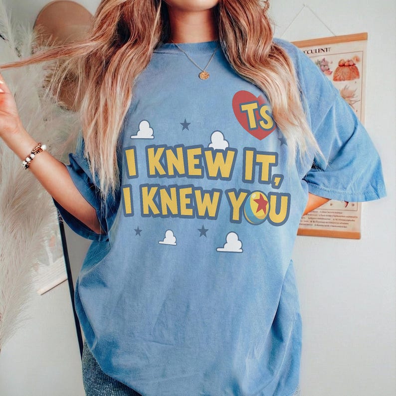Op de afbeelding: Een lichtblauw t-shirt met korte mouwen voorzien van een rood hartmotief met de letters TS, de tekst I KNEW IT, I KNEW YOU in het geel, en kleine witte wolkjes en donkergrijze sterren op de voorzijde van het kledingstuk.