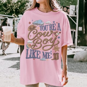 K&ouml;nnte beinhalten: Ein hellrosa T-Shirt mit kurzen &Auml;rmeln und einem Grafikdruck mit dem blauen Text "YOU'RE A COWBOY LIKE ME". Das Design zeigt einen braunen Cowboyhut, ein Lasso, eine Rakete, einen Sheriffstern, Cowboystiefel, Wolken und kleine Sterne.