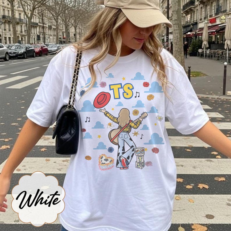 May include: A white t-shirt featuring a graphic print of a person with long hair playing a guitar, wearing a yellow plaid shirt and cow-print pants. The design includes blue clouds, a red hat, musical notes, and blocks with the letters TS.