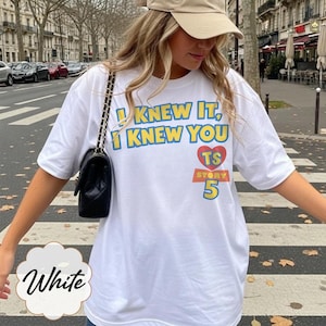 May include: A white short-sleeved t-shirt featuring the blue text I KNEW IT, I KNEW YOU above a red heart containing the letters TS, with the word STORY and the number 5 printed below in yellow and blue, paired with a black quilted shoulder bag.