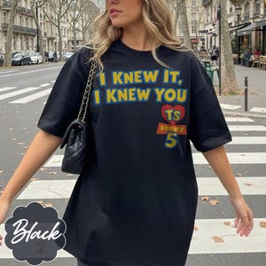 May include: A black short-sleeved t-shirt featuring the yellow text I KNEW IT, I KNEW YOU, a red heart with the letters TS, and the word STORY above the number 5 in yellow and blue. A black quilted shoulder bag with a chain strap is also visible.