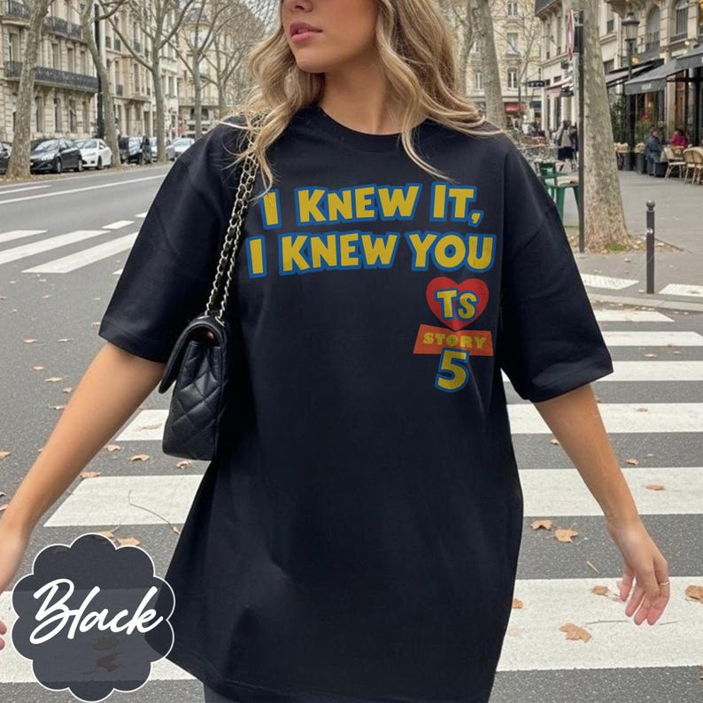 May include: A black short-sleeved t-shirt featuring the yellow text I KNEW IT, I KNEW YOU, a red heart with the letters TS, and the word STORY above the number 5 in yellow and blue. A black quilted shoulder bag with a chain strap is also visible.