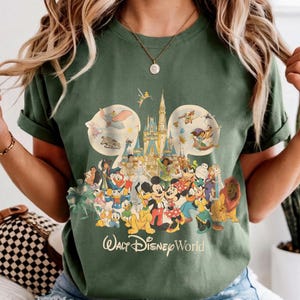 May include: An olive green crew neck t-shirt featuring a large graphic print of the Walt Disney World castle and various animated characters arranged in a Mickey Mouse ear silhouette, with the text Walt Disney World printed below the design.