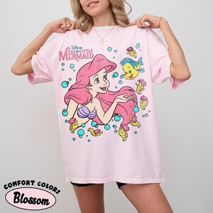 Puede incluir: Una camiseta rosa claro con un gr&aacute;fico de Ariel de La Sirenita de Disney, junto a Flounder y varios caballitos de mar rodeados de burbujas turquesas. El texto Disney The Little Mermaid est&aacute; impreso en una fuente estilizada sobre el personaje.