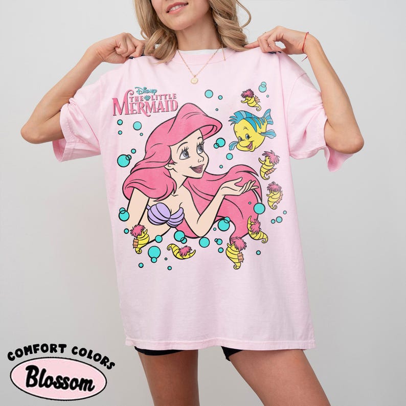 Puede incluir: Una camiseta rosa claro con un gr&aacute;fico de Ariel de La Sirenita de Disney, junto a Flounder y varios caballitos de mar rodeados de burbujas turquesas. El texto Disney The Little Mermaid est&aacute; impreso en una fuente estilizada sobre el personaje.
